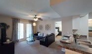 This single-floor residence offers a well-designed open-concept layout that seamlessly connects the main living areas. The living room is a welcoming focal point, featuring plush carpeted flooring, a ceiling fan with integrated lighting, and a textured ceiling. A large sectional sofa anchors the space, complemented by a sleek glass coffee table and decorative wall art. Natural light pours in through French doors dressed with elegant drapes, creating a bright and airy atmosphere. Adjacent to the living area, a dining space is visible with dark granite-style countertop seating, offering a casual yet functional area for meals. Decorative pendant lighting adds a stylish touch overhead. The bedroom, visible through an open doorway from the living room, features continued carpeted flooring, a ceiling fan, and a tasteful dark upholstered headboard, conveying a calm and restful retreat. The neutral color palette of warm beige and white tones throughout the space creates a cohesive and inviting ambiance. The thoughtful layout and connected flow between rooms make this residence both practical and comfortable for daily living.