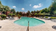 Located at 2323 Long Reach Dr in Sugar Land, Texas, this outdoor amenity space showcases a thoughtfully designed resort-style environment set against a backdrop of classic red brick multi-story buildings. The centerpiece of the grounds is a generously sized swimming pool with a smooth, curved design, complemented by a raised spa/hot tub area accessible via brick-accented steps. The pool deck is finished in warm-toned stone tile and furnished with striped lounge chairs, freestanding market umbrellas, and large decorative clay planters filled with vibrant seasonal flowers. Tall Italian cypress trees, soaring palm trees, and mature shade trees frame the pool area, creating a lush, resort-like atmosphere. Surrounding the pool, manicured landscape beds feature ornamental grasses, sculpted hedges, and colorful shrubs, all maintained to a high standard. Broad, well-lit concrete pathways wind through the grounds, connecting the pool area to residential buildings and green lawn spaces enclosed by wrought iron fencing. The buildings themselves feature arched entryways, dormer windows, and private balconies, contributing to a cohesive architectural character throughout the community. The overall ambiance is one of a well-maintained, upscale outdoor retreat in the heart of Sugar Land.