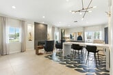 Located at 2235 Treehouse Ln in Corona, California, this single-floor space offers a cohesive, contemporary design that maximizes its footprint through an open, flowing layout. Large-format light-toned tile flooring extends throughout, lending a clean, airy feel to the entire area.

The main living area is anchored by a striking floor-to-ceiling textured tile fireplace surround, flanked by built-in display niches that add architectural depth. A seating arrangement featuring plush blue accent chairs and a comfortable gray sofa centers around a round wooden coffee table, all set atop a bold blue-and-white geometric area rug that provides a vivid focal point.

A sleek white marble-topped bar counter with black bar stools serves as a natural divider, defining a more functional gathering zone. An eye-catching multi-arm brass and black pendant light fixture overhead adds an artistic, modern touch. A warm wood slat partition wall creates a subtle separation toward the back of the space, adding texture and visual interest.

Multiple windows dressed with sheer linen curtains invite abundant natural light, while recessed lighting ensures consistent illumination throughout. The walls are adorned with large-scale abstract artwork in teal, gold, and blue tones, reinforcing the contemporary aesthetic. The overall ambiance is bright, polished, and welcoming.