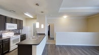 This single-story residence in Krum, Texas, offers an open-concept design that seamlessly connects the kitchen and living areas. The kitchen is a functional centerpiece, featuring a peninsula with a dark granite-style countertop, an integrated sink, and dark espresso cabinetry that provides ample storage. A white refrigerator anchors one end of the kitchen, while pendant lighting fixtures hang above the peninsula, adding both task lighting and a modern aesthetic touch. Recessed and flush-mount ceiling lights ensure the space is well-illuminated throughout.

The flooring consists of durable hardwood-style planks in a warm, medium-toned finish that run continuously through the kitchen and into the adjoining living area, creating visual cohesion across the open layout. A half-wall partition subtly defines the transition between the kitchen and living space while maintaining the open feel. Crown molding details along the upper walls add a refined architectural touch.

A hallway visible from the main living area leads to additional rooms, suggesting a well-organized floor plan. The neutral wall tones and clean white trim throughout contribute to a bright, welcoming ambiance well-suited to everyday living.