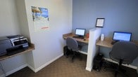 This single-floor office space is designed with functionality and productivity in mind. The workspace features built-in workstation counters with a stone-like laminate surface, providing ample desk space for computer setups. The workstations are divided by low partition walls, creating semi-private areas that balance collaboration with focused individual work. Each station is equipped with a desktop computer and accompanying peripherals, making the space ready for immediate use. The walls are painted in a calming blue-gray tone, contributing to a focused and professional atmosphere. Carpeted flooring in a neutral tone adds a layer of comfort underfoot while maintaining a tidy, business-appropriate aesthetic. The space is neatly organized, with tower PCs tucked beneath the counters to maximize usable desk area. Wall-mounted frames and decor add a touch of personality without distracting from the workspace's core purpose. Overall, this office space offers a practical and well-organized environment suited for administrative or clerical work, with a clean design that keeps the emphasis on efficiency and ease of use.