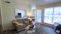 This single-floor residence offers a thoughtfully designed layout that balances comfort and style throughout. The living area serves as the heart of the space, featuring a textured ceiling and light, neutral-toned walls that create a warm and inviting atmosphere. A large decorative mirror with a geometric frame anchors the room, enhancing the sense of openness and reflecting natural light throughout. The space is filled with natural light streaming in through a full-length glass door and a sizable window fitted with blinds, offering a view of the outdoors and providing direct access to a private balcony or patio. The living area is thoughtfully arranged with a plush sofa, a patterned area rug, and a glass-top coffee table, contributing to an elegant yet approachable aesthetic. The room flows naturally into a dining area visible in the background, suggesting an open-concept design that supports both everyday living and entertaining. Dark hardwood-style flooring runs through the connected spaces, adding a rich contrast to the light walls and furnishings. Overall, this residence presents a cohesive and polished interior with an emphasis on natural light, functional flow, and refined design details.
