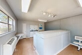 Located at 12703 Jones Road in Houston, Texas, this single-room community laundry facility offers a practical and organized space designed for shared residential use. The interior features vertical panel wall cladding painted in a neutral gray tone, complemented by a light-colored tile floor throughout. Illumination is provided by a combination of overhead fluorescent fixtures, a skylight that introduces natural light, and adjustable track lighting, ensuring the space is well-lit at all times. A row of top-load washing machines lines one wall, accompanied by stacked dryer units, all equipped with CSC GO app-enabled payment technology that supports tap-to-pay and mobile payments for added convenience. A wall-mounted air conditioning unit provides climate control, keeping the room comfortable year-round. Built-in laminate folding counters are positioned along the walls, offering ample surface area for sorting and folding laundry. A freestanding bench provides a designated waiting area for users. The space also includes a wall-mounted fire extinguisher box, reflecting attention to safety. A window along one wall allows views of the surrounding outdoor greenery, adding a touch of openness to the otherwise utilitarian space. The facility's straightforward, functional layout maximizes usability within its compact footprint.