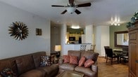 This single-floor space offers an open-concept layout that seamlessly integrates the living, dining, and kitchen areas into one cohesive and functional environment. The living room serves as the heart of the space, featuring rich brown leather sofas adorned with decorative pillows in warm earth tones, centered around a ceiling fan that provides both style and comfort. Warm wood-style flooring flows throughout, adding a sense of continuity and warmth to the interior. The kitchen is positioned just beyond the living area and is highlighted by light wood cabinetry, a black appliance suite including a microwave and refrigerator, and a raised breakfast bar that doubles as a casual dining counter. Bar-style seating at the counter provides a convenient spot for quick meals or conversation. A dedicated dining area is visible nearby, offering additional seating for more formal meals. Arched architectural details frame the transition between spaces, adding a subtle design element that enhances the overall character of the interior. The layout is thoughtfully designed to maximize both functionality and flow, making this space well-suited for everyday living and entertaining alike.