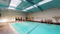 This indoor pool facility is a well-appointed aquatic space designed for both recreation and relaxation. The centerpiece is a generously sized lap pool with clear, turquoise water, complemented by a separate hot tub located at one end of the facility. The soaring ceiling is a defining architectural feature, finished in a signature teal blue and supported by exposed wooden beams that lend the space a warm, resort-like character. A series of skylights runs along the ceiling's length, flooding the interior with abundant natural light throughout the day. Louvered and mesh-screened windows line the perimeter walls, providing additional ventilation and soft, diffused daylight. The surrounding deck is finished in large-format concrete tiles and furnished with multiple sling-style lounge chairs, offering ample space for relaxation poolside. Potted indoor trees are placed throughout the deck area, adding a touch of greenery and enhancing the welcoming atmosphere. A separate hot tub area is accessible via entry doors and includes frosted glass panels and posted pool rules for user guidance. The facility also includes utility access points and safety equipment, reflecting a thoughtfully maintained and fully functional aquatic environment.