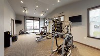 Located at 1608 East South Riverton Avenue in Spokane, Washington, this single-floor fitness center is thoughtfully designed to support a comprehensive workout experience. The space features vaulted ceilings with recessed lighting that create an open, airy atmosphere, while neutral gray walls and textured carpet flooring lend a clean, modern aesthetic throughout.

The exercise area is well-equipped with a variety of Matrix-brand cardio machines, including treadmills, upright and recumbent stationary bikes, an elliptical, and a rowing machine. A dedicated Training Zone multi-functional rack provides storage for dumbbells, resistance bands, and TRX suspension training straps, making strength and functional training easily accessible. A wall-mounted flat-screen television adds convenience for entertainment during workouts.

A standout architectural feature is the full-width glass-panel garage door, which opens directly onto an outdoor patio area furnished with seating — allowing for natural ventilation and seamless indoor-outdoor connectivity. Multiple windows with blinds provide additional natural light throughout the space.

The facility also includes a restroom, accessible via a clearly marked door, and a small utility area. The entryway features a standard door with a transom window, offering a welcoming point of access. Overall, the fitness center combines practical amenities with a well-appointed design suited for daily exercise.