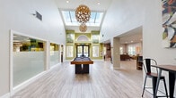 Located at 2053 NE 106th Ave in Hillsboro, Oregon, this single-floor space offers a generous, open-concept layout that seamlessly connects the kitchen, dining, and living areas. The kitchen is outfitted with crisp white cabinetry, stainless steel appliances including a range, microwave, dishwasher, and French door refrigerator, all set atop light wood-look flooring. A central island with pendant lighting and leather-style bar seating provides both functionality and a stylish focal point for casual dining. The open layout transitions into a bright, expansive living room anchored by a dramatic floor-to-ceiling stone fireplace with a rustic wood mantle. Vaulted ceilings with recessed lighting enhance the sense of openness, while multiple windows allow abundant natural light to fill the space. The living area features plush carpeting and ample room for comfortable seating arrangements. An additional area with light wood flooring accommodates a full-size pool table, adding a recreational dimension to the space. Accent walls in soft green complement the neutral palette throughout, and colorful artwork contributes to the welcoming, contemporary ambiance of this well-appointed Hillsboro property.