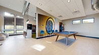 Located at 5 Rivers Run Dr in Schenectady, NY, this single-floor amenity center offers a generous and thoughtfully designed space for fitness, recreation, and socializing. The facility is organized into distinct zones, each serving a specific purpose while maintaining a cohesive modern aesthetic throughout.

The fitness center is a standout feature, bathed in natural light streaming through floor-to-ceiling windows that frame views of surrounding greenery. Motivational text lines the walls above the windows, reinforcing the energetic atmosphere. The exercise area is equipped with a full array of cardio machines, including multiple treadmills and ellipticals, as well as strength-training equipment, free weights, and functional fitness stations. A blue and gray tile floor adds a bold visual accent to the space.

Adjacent to the fitness area, a spacious lounge and game room offers a social hub furnished with stylish leather seating, high-top tables, and pendant lighting. A dramatic full-wall mural serves as a focal point, complemented by a pool table, an electronic dart board arcade machine, and a wall-mounted flat-screen TV. Exposed ductwork adds an industrial touch to the contemporary design.

A dedicated indoor sports simulator room rounds out the amenities, featuring an artificial turf mat, impact-absorbing wall panels, a projector system, and a rack of sports equipment for a versatile interactive experience. Large glass entry doors and abundant windows ensure the entire space feels open, bright, and connected to the outdoors.