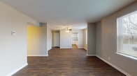This single-story residence offers a well-organized, open layout with consistent dark hardwood-style flooring running throughout the main living areas and bedrooms, creating a cohesive and grounded aesthetic. Neutral gray and beige tones on the walls complement the flooring, lending a clean, understated character to each room. Textured ceilings are present throughout, and ceiling fans with integrated lighting fixtures are installed in the living areas and bedrooms, providing both airflow and ambient light. The spacious open living area flows toward a dining space adjacent to the kitchen, which features white cabinetry, generous counter space with dark laminate countertops, a double stainless steel sink, and a window overlooking the outdoors. Built-in shelving is visible near the kitchen, offering additional storage or display space. The bedrooms are generously sized, each equipped with a ceiling fan and access to natural light via sliding glass doors dressed with vertical blinds that open to outdoor views. Two full bathrooms serve the residence; one features a white vanity with a large mirror, tiled tub-surround, and classic basket-weave tile flooring, while the second is a more compact bath with a tiled tub-surround and hardwood-style flooring. The overall layout is functional and move-in ready.