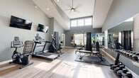 Located at 9060 Heirloom Ln in Reynoldsburg, Ohio, this well-equipped fitness center offers a comprehensive workout environment within a single, open-plan exercise room. The space is defined by its striking vaulted ceilings, which create an expansive, airy atmosphere and are complemented by clerestory windows that flood the room with natural light. Light wood-tone flooring runs throughout, contributing to a clean and modern aesthetic.

The cardio zone is equipped with a treadmill, elliptical trainer, and stationary bike, all positioned beneath wall-mounted flat-screen televisions for entertainment during workouts. A multi-function cable machine provides options for strength training, while a dedicated free weights area features a full dumbbell rack, adjustable weight bench, and an assortment of exercise balls and step platforms for functional and flexibility training. A fold-out exercise mat mounted to the wall offers additional versatility.

Full-length mirrors along one wall visually expand the space and support form-focused training. A ceiling fan overhead aids air circulation, and glass-paned doors connect the gym to an adjacent common area, enhancing the sense of openness. The cool gray wall tones and organized layout combine to create a focused yet welcoming fitness environment.