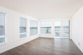 This well-designed single-floor residence offers a clean, contemporary aesthetic throughout, characterized by crisp white walls, light-toned luxury vinyl plank flooring in the main living areas, and an abundance of natural light streaming through multiple large windows fitted with horizontal blinds.

The open-concept living and dining area is generously proportioned, with light wood-look flooring that creates a warm, airy atmosphere. A bank of wide windows frames scenic outdoor views of the surrounding cityscape and water, filling the space with natural light. The kitchen flows seamlessly from the living area and is outfitted with flat-front white cabinetry, dark countertops, a stainless steel sink, and a full suite of stainless steel appliances including a range, over-the-range microwave, dishwasher, and refrigerator with ice dispenser — offering both functionality and a modern aesthetic.

The bedrooms feature soft wall-to-wall carpeting and windows with outdoor views. At least one bedroom includes a dedicated walk-in closet with built-in shelving, providing ample storage. The bathroom is thoughtfully designed with accessibility in mind, featuring grab bars, a spacious open layout, wood-look flooring, and a white vanity with a dark countertop. The unit's cohesive neutral palette and modern finishes create a bright, move-in-ready living environment.