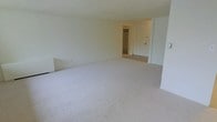 This single-floor unit features a spacious, open living area finished with light-toned wall-to-wall carpeting that lends a clean and neutral aesthetic throughout. The walls are painted in a crisp white, contributing to a bright and airy atmosphere. Toward the back of the room, an open doorway leads to a closet equipped with built-in shelving, offering practical storage options. Adjacent to the closet is an entry door, providing a clear view of the layout's functional flow. The ceilings are standard height, maintaining a comfortable and proportionate sense of scale within the room. The overall design is straightforward and versatile, with a blank-canvas quality that easily accommodates a variety of furnishing arrangements and décor styles. The absence of clutter highlights the room's generous open floor plan, making it feel expansive and easy to configure. The neutral palette throughout creates a cohesive and understated ambiance well-suited for everyday living.