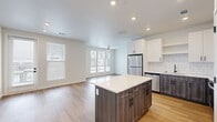 Located in Denver's vibrant Central Park neighborhood, this well-appointed one-bedroom, one-bathroom residence offers a thoughtfully designed single-floor layout. The open-concept kitchen and living area serves as the heart of the home, anchored by a substantial kitchen island with a wood-grain finish and white quartz countertop. The kitchen is outfitted with crisp white upper cabinetry, contrasting dark lower cabinets, a classic subway tile backsplash, and a full suite of stainless steel appliances including a refrigerator, range, microwave, and dishwasher. Recessed lighting illuminates the space throughout. The living area flows naturally from the kitchen and is brightened by a bank of large windows and a ceiling fan, creating an airy, comfortable atmosphere. Light hardwood flooring runs continuously throughout the entire residence, lending warmth and visual cohesion. The bedroom is generously proportioned, featuring multiple windows on two walls that invite abundant natural light and neighborhood views, along with a ceiling fan for comfort. A spacious walk-in closet with built-in wire shelving and hanging rods provides ample storage. The bathroom is finished with a floating vanity with quartz countertop, black hardware accents, a large mirror, and stylish overhead lighting, all carried through with the same hardwood flooring.