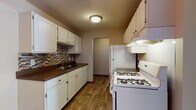This single-floor residence offers a practical and comfortable layout across several well-defined living spaces. The galley-style kitchen is a functional highlight, featuring white cabinetry with ample upper and lower storage, dark laminate countertops, and a mosaic tile backsplash in warm earth tones. White appliances include a gas range and a full-size refrigerator, complemented by an under-cabinet range hood and a built-in dishwasher. Wood-look flooring runs throughout the kitchen, adding warmth to the space. The living room is generously sized, finished with plush carpeting, a textured ceiling, and a large sliding glass door that opens to a private balcony, inviting natural light and outdoor views. The bedroom offers soft carpeting, a ceiling fan with light fixture, a closet, and a broad window framing exterior views. An additional room with carpeting and a ceiling-mount light fixture provides flexible use as a second bedroom or den. The bathroom includes a full soaking tub with white tile surround, a toilet, and a vanity with countertop, all framed by warm wood-toned door trim. Throughout the unit, baseboard heating, neutral wall tones, and wood-accented trim details contribute to a cohesive and livable aesthetic.