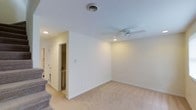 This two-floor space is designed with a clean, functional aesthetic throughout. The main living area is finished with plush wall-to-wall carpeting in a neutral beige tone, contributing to a warm and comfortable atmosphere. White walls and white trim create a bright, airy feel, while recessed lighting and a ceiling fan with integrated lighting provide both ambient and directed illumination. A centrally positioned ceiling vent ensures efficient climate control throughout the room. Adjacent to the main living area is a tiled bathroom, visible through an open doorway, offering convenient access. The staircase, partially visible in the corner, features carpeted treads that match the main floor, connecting seamlessly to the second level. The overall layout is open and uncluttered, allowing for flexible furniture arrangements. The neutral color palette and simple finishes throughout give the space a versatile, move-in-ready quality suitable for a variety of uses and personal styles.