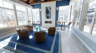 This single-floor space makes a strong first impression with its dramatic, high ceilings accented by exposed wood beams, creating a sense of openness and architectural character. The interior is defined by a bold navy and white color palette that carries throughout the room, from the deep blue accent walls to the large area rug with geometric patterning anchoring the central seating arrangement.

A linear gas fireplace serves as the room's focal point, framed by a crisp white surround and flanked by a striking abstract artwork above. Surrounding the fireplace, generous windows flood the space with natural light, offering views of the exterior surroundings and enhancing the airy atmosphere.

The seating is thoughtfully arranged to encourage conversation, featuring plush tan lounge chairs gathered around a glass-top coffee table with teal decorative accents. Along the perimeter, high-top bar tables with swivel stools provide additional seating options. The flooring transitions from light gray tile at the edges to the plush area rug at the center, adding warmth and texture. Overall, the space balances a contemporary aesthetic with inviting comfort.