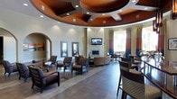 This single-floor communal space makes an immediate impression with its dramatic coffered ceiling, finished in deep burnt orange and crisp white geometric patterns with recessed lighting throughout. The open-concept layout is anchored by a central lounge area featuring a sleek fireplace with a decorative tile surround and a wall-mounted flat-screen television, around which curved and upholstered seating arrangements invite comfortable gatherings. Large-format tile flooring extends across the space, grounding the room with a clean, contemporary aesthetic. Multiple distinct seating zones are thoughtfully arranged throughout — from cushioned armchairs and low coffee tables to bar-height seating along the perimeter — offering flexibility for both casual conversation and independent use. Arched doorways add a touch of architectural elegance, creating visual continuity between areas. Tall windows dressed with bold striped draping in complementary rust, navy, and cream tones allow natural light to filter in while enhancing the room's warm color palette. The overall ambiance is sophisticated yet welcoming, blending modern design elements with inviting furnishings to create a versatile, well-appointed communal lounge environment.