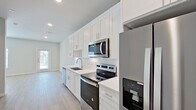 This well-appointed single-floor space showcases a thoughtful, modern design with a consistent neutral palette and clean finishes throughout. The kitchen is a standout feature, equipped with floor-to-ceiling white shaker cabinetry, white quartz countertops, a classic subway tile backsplash, and a full suite of stainless steel appliances including a French door refrigerator with water dispenser, electric range, over-the-range microwave, and dishwasher. Recessed lighting and a window at the end of the kitchen allow natural light to fill the space. Light hardwood-style flooring runs throughout the unit, providing a warm and cohesive feel underfoot. The entryway is clean and functional, with a smart-lock front door flanked by additional interior doors offering convenient storage or utility access. The living area is bright and open, benefiting from a ceiling fan and a large window that invites natural light. A second room features a glass exterior door, offering direct access to the outside and additional natural illumination. The bathroom is finished with neutral tile flooring, a soaking tub with tiled surround, a white vanity, and clean, simple fixtures. Overall, the space presents a fresh, move-in-ready environment with modern appointments and a light, airy ambiance.