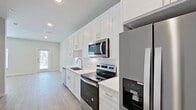This well-appointed single-floor space showcases a thoughtful, modern design with a consistent neutral palette and clean finishes throughout. The kitchen is a standout feature, equipped with floor-to-ceiling white shaker cabinetry, white quartz countertops, a classic subway tile backsplash, and a full suite of stainless steel appliances including a French door refrigerator with water dispenser, electric range, over-the-range microwave, and dishwasher. Recessed lighting and a window at the end of the kitchen allow natural light to fill the space. Light hardwood-style flooring runs throughout the unit, providing a warm and cohesive feel underfoot. The entryway is clean and functional, with a smart-lock front door flanked by additional interior doors offering convenient storage or utility access. The living area is bright and open, benefiting from a ceiling fan and a large window that invites natural light. A second room features a glass exterior door, offering direct access to the outside and additional natural illumination. The bathroom is finished with neutral tile flooring, a soaking tub with tiled surround, a white vanity, and clean, simple fixtures. Overall, the space presents a fresh, move-in-ready environment with modern appointments and a light, airy ambiance.