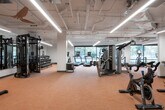 This fitness center along the Interurban Trail in Shoreline, WA offers a comprehensively equipped, single-floor facility designed to support a wide range of workout routines. The space is organized into distinct functional zones, beginning with a generously sized main floor housing an array of cardio equipment, including Matrix treadmills, ellipticals, rowing machines, and cycling bikes positioned along floor-to-ceiling windows. These large-format windows invite abundant natural light and frame views of the surrounding Pacific Northwest greenery, creating an engaging environment for cardio workouts. A dedicated strength and functional training section includes cable machines, weight racks, and benches distributed throughout the open floor plan, supported by exposed ductwork ceilings that lend an industrial character to the space. A separate studio area features full-length mirrored walls, wall-mounted flat-screen televisions, and smaller display screens, alongside accessories such as TRX straps, resistance tools, kettlebells, and exercise mats, making it suitable for group training or individual functional workouts. Warm-toned carpet flooring runs throughout the facility, and linear LED lighting provides consistent, bright illumination. The overall layout is open and well-organized, offering a professional and functional fitness environment.