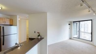 This single-floor space features a clean, minimalist design with an open-concept layout that connects the kitchen and living areas seamlessly. A dark-toned countertop acts as a natural divider between the kitchen and the adjoining living space, providing both functional workspace and a subtle visual boundary. The living area is finished with neutral-toned carpet, contributing to a warm and understated atmosphere throughout.

White walls and a textured ceiling maintain a bright, airy feel across the space. A large window at the far end of the living area allows natural light to filter in, enhancing the sense of openness. A hallway visible from the main area leads to additional rooms, including what appears to be a bathroom with light-toned finishes.

The overall design is practical and uncluttered, with a neutral color palette that provides a versatile backdrop suitable for a range of furnishing styles. The layout makes efficient use of the available space, with clear sightlines from the kitchen counter through to the living area, offering a cohesive and functional living environment.