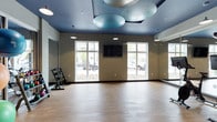 This contemporary fitness studio is thoughtfully designed to support a variety of workout activities. The space is anchored by a smooth hardwood-style floor that provides an open, flexible area ideal for group exercise, stretching, or free movement. Large white-framed windows line the exterior wall, allowing abundant natural light to fill the room while offering views of the surrounding outdoor environment.

A standout architectural feature is the bold blue ceiling, which adds a dynamic visual element to the space. Suspended from the ceiling are large, rounded acoustic panels in a muted steel-blue tone, designed to manage sound within the room — a practical and stylish detail for an active fitness environment. Recessed and pendant-style lighting complements the natural light, ensuring the space remains well-lit at all times.

A wall-mounted flat-screen television is positioned prominently for easy viewing during workouts. A dumbbell rack along one wall provides convenient access to free weights, while large mirrors enhance the sense of openness and allow users to monitor their form. The neutral-toned walls contrast cleanly with the blue ceiling, creating a focused yet energizing atmosphere well-suited for an active fitness facility.