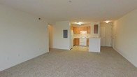 This single-floor space features an open-concept design that seamlessly connects the living area to the kitchen. The living area is finished with neutral-toned carpet, creating a comfortable and versatile space that can accommodate a variety of furniture arrangements. White walls throughout contribute to a bright, clean aesthetic.

The kitchen is positioned at the rear of the open layout and is defined by a peninsula countertop that serves as a natural divider between the two areas. Honey-toned wood cabinetry lines the kitchen walls, providing ample storage, while white appliances — including a refrigerator and electric range — are neatly integrated beneath the overhead cabinets. A ceiling-mounted light fixture illuminates the kitchen workspace. The kitchen floor transitions to a light-colored hard surface, offering a practical contrast to the carpeted living space.

A structural column with an electrical panel is visible along one wall, and a hallway leading to additional areas of the unit can be seen to the left. The overall ambiance is functional and straightforward, with a neutral palette that offers flexibility for personalization.