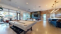 Located at 3400 E Southern Ave in Phoenix, Arizona, this single-floor community clubhouse offers a generously sized, open-concept layout designed for both social engagement and relaxed lounging. The space features polished concrete floors throughout, lending a sleek, industrial aesthetic that is warmed by a striking reclaimed wood accent wall serving as a central design focal point.

The main lounge area is anchored by a large wall-mounted flat-screen television, complemented by a curated seating arrangement of modern sofas and leather accent chairs. A full-size billiards table provides a dedicated recreational zone, while expansive floor-to-ceiling steel-framed windows and glass doors flood the interior with natural light and offer views of the outdoor amenity areas.

A stylish kitchenette features a navy tile-clad island with bar seating, a coffee station, under-counter refrigerator, and industrial pendant lighting overhead. The space also includes glassed-in private office or conference rooms, offering a quieter setting for focused work or meetings. Geometric wall art and lush indoor plants add curated character throughout. The overall ambiance is contemporary and welcoming, blending functionality with upscale residential design.