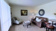 This single-floor residential space offers a comfortable and welcoming atmosphere, centered around a thoughtfully arranged living area. The living room features plush wall-to-wall carpeting in a warm neutral tone, providing a soft and inviting foundation for the space. Smooth, light-colored walls are complemented by a textured ceiling, lending the room a classic, homey character. The seating arrangement includes a rich brown leather loveseat adorned with decorative pillows and a traditional upholstered accent chair, both positioned around a stylish tufted ottoman that serves as a central coffee table. Subtle decorative touches — including a large wall clock, a framed mirror, and a table lamp — add personality and warmth to the room without overwhelming the clean, understated aesthetic. A glimpse into the adjacent kitchen is visible through the mirror's reflection, hinting at a connected, open layout between the living and culinary spaces. The kitchen appears bright with light cabinetry. Overall, the space presents a cozy, move-in-ready environment with a cohesive neutral palette, practical layout, and warm, classic design elements that create an immediately livable and comfortable home.