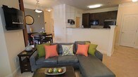 This single-floor residence features an open-concept layout that seamlessly connects the living, dining, and kitchen areas into one cohesive, functional space. The living area is anchored by a large sectional sofa accented with colorful throw pillows, paired with a dark coffee table that adds contrast to the light-toned flooring and neutral walls. Adjacent to the living space, a compact dining area accommodates a round dark-wood table with matching chairs, ideal for intimate meals. A pendant light fixture overhead adds a touch of warmth to the dining nook. The kitchen, visible from both the living and dining areas, features dark cabinetry, granite-style countertops, and black appliances, including a built-in microwave. A raised breakfast bar serves as a natural divider between the kitchen and the main living space, enhancing the open feel while offering additional seating or serving options. Flush-mounted ceiling lighting throughout ensures consistent illumination. Neutral wall tones and light flooring create a bright, cohesive aesthetic, while the open sightlines across all areas contribute to a sense of spaciousness throughout the home.