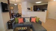This single-floor residence features an open-concept layout that seamlessly connects the living, dining, and kitchen areas into one cohesive, functional space. The living area is anchored by a large sectional sofa accented with colorful throw pillows, paired with a dark coffee table that adds contrast to the light-toned flooring and neutral walls. Adjacent to the living space, a compact dining area accommodates a round dark-wood table with matching chairs, ideal for intimate meals. A pendant light fixture overhead adds a touch of warmth to the dining nook. The kitchen, visible from both the living and dining areas, features dark cabinetry, granite-style countertops, and black appliances, including a built-in microwave. A raised breakfast bar serves as a natural divider between the kitchen and the main living space, enhancing the open feel while offering additional seating or serving options. Flush-mounted ceiling lighting throughout ensures consistent illumination. Neutral wall tones and light flooring create a bright, cohesive aesthetic, while the open sightlines across all areas contribute to a sense of spaciousness throughout the home.