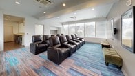 This single-floor amenity space is thoughtfully designed to serve multiple functions, combining entertainment, collaboration, and productivity in a contemporary setting. The signature feature is a generously sized screening room outfitted with rows of black leather power recliner chairs arranged in a tiered-style layout, offering comfortable viewing of the wall-mounted flat-screen television. The room is accented by bold yellow and gray wall colors, recessed ceiling lighting, and distinctive multi-toned blue and gray plank flooring that carries a striking visual character throughout the space. Large floor-to-ceiling windows along one wall flood the screening area with natural light and provide views of an outdoor deck and surrounding neighborhood. A built-in bench with open storage sits beneath the windows, complemented by a small side table. Adjacent to the screening room is a compact private meeting room featuring a round white table, modern ergonomic chairs, and a glass panel wall element, well-suited for small group discussions. The business center area features warm wood-tone flooring, a long white countertop workstation equipped with a computer and printer set against a vibrant green accent wall, and a bar-height counter with green and chrome stools along the opposite side, ideal for focused work or casual collaboration.