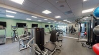 Located along River Road in North Little Rock, Arkansas, this single-floor fitness center is designed to support a comprehensive workout experience. The space is finished with durable rubber flooring throughout, a drop ceiling with recessed panel lighting, and sage green accent walls that lend the facility a clean, modern aesthetic. Full-length mirrors line multiple walls, enhancing the sense of openness and allowing users to monitor their form during exercise.

The cardio zone features a row of treadmills, elliptical trainers, and upright cycling bikes, each equipped with individual display screens. Multiple flat-screen televisions are wall-mounted above the cardio equipment for added entertainment. Adjacent to this area, a dedicated cycling section houses spin bikes ideal for high-intensity interval training.

The strength training section is well-stocked with a full rack of dumbbells, adjustable benches, and a cable resistance machine for targeted muscle training. A functional training corner includes medicine balls of varying weights, stability balls, and additional accessories stored on organized racks. A multi-use cable and weight machine anchors the strength zone.

Natural light filters into the space through large windows and a glass-paneled exterior wall, offering views of the surrounding outdoor area. A vending machine provides convenient on-site refreshments, and the overall layout ensures ample space for movement between equipment zones.