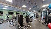 Located along River Road in North Little Rock, Arkansas, this single-floor fitness center is designed to support a comprehensive workout experience. The space is finished with durable rubber flooring throughout, a drop ceiling with recessed panel lighting, and sage green accent walls that lend the facility a clean, modern aesthetic. Full-length mirrors line multiple walls, enhancing the sense of openness and allowing users to monitor their form during exercise.

The cardio zone features a row of treadmills, elliptical trainers, and upright cycling bikes, each equipped with individual display screens. Multiple flat-screen televisions are wall-mounted above the cardio equipment for added entertainment. Adjacent to this area, a dedicated cycling section houses spin bikes ideal for high-intensity interval training.

The strength training section is well-stocked with a full rack of dumbbells, adjustable benches, and a cable resistance machine for targeted muscle training. A functional training corner includes medicine balls of varying weights, stability balls, and additional accessories stored on organized racks. A multi-use cable and weight machine anchors the strength zone.

Natural light filters into the space through large windows and a glass-paneled exterior wall, offering views of the surrounding outdoor area. A vending machine provides convenient on-site refreshments, and the overall layout ensures ample space for movement between equipment zones.