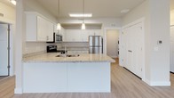 This single-floor residence offers a thoughtfully designed open-concept layout that seamlessly connects the kitchen, dining, and living areas. Light wood-look flooring flows throughout the main living spaces, complemented by neutral-toned walls and abundant natural light streaming through multiple windows fitted with white blinds. Ceiling fans are installed in both the living area and bedroom, enhancing comfort and airflow.

The kitchen is a standout feature, showcasing white shaker-style cabinetry, light granite countertops, and a large peninsula with an undermount sink — ideal for both meal preparation and casual dining. A full suite of stainless steel appliances, including a refrigerator, range, microwave, and dishwasher, completes the space. Pendant lighting above the peninsula adds a modern touch.

The bedroom is generously sized with plush carpeting, multiple windows, and a ceiling fan. It is complemented by a walk-in closet outfitted with dual hanging rods and a built-in shelving tower for organized storage.

The full bathroom features white cabinetry, granite countertops, an undermount sink, a large mirror, and a soaking tub with shower combination. A dedicated laundry area houses a stacked front-loading washer and dryer unit for added convenience.