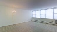 This single-floor space presents a clean, open living area finished with wall-to-wall neutral carpeting that adds warmth underfoot. The room is painted in crisp white throughout, creating a bright and airy atmosphere. A classic candelabra-style chandelier serves as a decorative focal point overhead, adding a touch of traditional elegance to the otherwise minimalist setting. Large windows along one wall allow generous natural light to flood the interior, enhancing the sense of openness. A baseboard heating unit runs beneath the windows, offering a practical and efficient heating solution. The smooth white walls and simple trim provide a neutral backdrop well-suited for a variety of furnishing styles. The space feels open and versatile, functioning well as a living or dining area. Overall, the room conveys a straightforward, functional design with classic detailing.