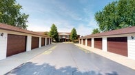 This well-maintained residential complex presents a functional and orderly layout centered around a wide paved courtyard. Flanking both sides of the approach are rows of individual garage units, each fitted with brown overhead doors set against white-painted exteriors, providing generous covered parking and storage options for residents. The courtyard itself is clearly marked with yellow painted designations, ensuring organized vehicle flow throughout the property.

At the end of the courtyard stands a two-story brick building, featuring a traditional architectural style with a dark brown pitched roof, symmetrical facade, and a welcoming entrance framed by columns. A covered entryway adds a classic touch to the exterior. Mature deciduous trees are thoughtfully placed throughout the property, offering shade, visual interest, and a sense of established character. The landscaping contributes to a pleasant, park-like atmosphere around the building's entrance.

The overall setting conveys a practical yet comfortable community environment, balancing accessible parking infrastructure with attractive greenery and traditional brick construction. The open courtyard enhances the sense of space, giving the property an airy, accessible feel.