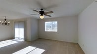 This single-story space presents a clean and functional layout centered around a well-lit main living area. The room features wall-to-wall carpet in a neutral beige tone, contributing to a warm and understated aesthetic throughout. Light-colored walls provide a versatile backdrop, making the space feel open and adaptable to a variety of interior styles.

A ceiling fan with an integrated light fixture is centrally mounted on the textured ceiling, offering both air circulation and ambient lighting. Natural light filters into the room through a window fitted with horizontal blinds, allowing for adjustable light control. A set of vertical blinds on an adjacent wall panel suggests the presence of a secondary window or sliding door, further enhancing the brightness of the space.

The room's layout is straightforward and spacious, with clean baseboards framing the perimeter. The overall ambiance is calm and neutral, offering a move-in-ready environment that is easy to personalize. The single-floor design ensures convenient accessibility throughout, making it a practical and comfortable living space.