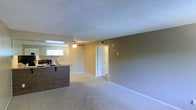 This single-story space features an open-concept layout that seamlessly connects the kitchen and living area. The living space is finished with wall-to-wall carpeting in a neutral tone, complemented by warm beige walls and white baseboards that contribute to a clean, cohesive aesthetic. A ceiling fan is centrally positioned overhead, providing both air circulation and ambient lighting to the main living area.

The kitchen is partially separated by a wood-toned countertop bar that serves as a natural divider between the two spaces while maintaining an open feel. A window near the kitchen area allows natural light to filter in, brightening the interior. The kitchen includes visible appliances and functional counter space.

A textured popcorn ceiling runs throughout the space, adding a traditional character to the interior. A white interior door is visible at the far end of the living area, leading to additional rooms or a hallway. A wall-mounted vent ensures climate control throughout. Overall, the space presents a functional, straightforward layout with practical finishes suited for comfortable everyday living.