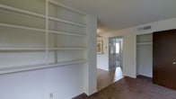 This single-story space showcases a straightforward, functional layout with a notable built-in shelving unit as a central design element. The floor-to-ceiling shelves are painted white and offer generous storage or display capacity, anchored by a structural column that subtly defines separate zones within the open layout. The floors are covered in neutral-toned carpet, contributing to a warm and quiet atmosphere throughout. A textured popcorn ceiling spans the space, consistent with a classic residential aesthetic. Looking down the central corridor, louvered double doors provide access to an adjoining room, while natural light filters in through a window visible at the far end, brightening the hallway. Framed wall art adds a touch of character to the passage. A floor vent is visible along the baseboard, indicating a standard forced-air climate system. The walls are finished in a clean white, keeping the interior feeling bright and neutral. Overall, the space offers a practical, move-in-ready environment with ample built-in storage and a connected, easy-to-navigate floor plan.