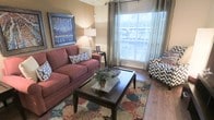 This single-floor space offers a warm and inviting atmosphere defined by thoughtful design and comfortable living. The living room serves as the heart of the space, showcasing rich hardwood flooring that complements the earthy, neutral wall tones throughout. A large window dressed with layered blinds and floor-length grommet curtains floods the room with natural light, creating an airy and open feel.

The living area is furnished with a plush sofa in a deep mauve tone, paired with a patterned accent chair in a bold geometric print, offering a pleasing contrast of styles. A dark wood coffee table anchors the seating arrangement over a vibrant, multi-colored area rug featuring intricate geometric patterns that add depth and character to the floor. Decorative throw pillows and a potted plant centerpiece on the coffee table contribute to the cozy, lived-in charm.

Crown molding details along the ceiling add an architectural elegance to the space, while a framed landscape painting on the wall introduces a pop of color and artistic flair. A classic table lamp provides warm ambient lighting to complement the natural light. Overall, the space strikes a balance between comfort and style, making it an ideal setting for relaxed, everyday living.