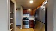 This single-floor unit offers a practical and well-appointed layout designed for comfortable everyday living. The kitchen is a functional highlight, featuring rich medium-toned wood cabinetry, stainless steel appliances including an electric range and a large refrigerator, stone-finish countertops, and a classic white subway tile backsplash. Tile flooring grounds the kitchen area, which flows seamlessly into the open living space finished with light hardwood-style flooring throughout.

The living area is open and versatile, with neutral gray walls, a drop tile ceiling, and white baseboards that provide a clean, cohesive aesthetic. A hallway connects the main living areas to the private spaces beyond, including a bedroom that benefits from a large window offering outdoor views and ample natural light, complementing the light hardwood flooring and neutral-toned walls.

The bathroom is neatly appointed with a white vanity, a medicine cabinet with lighted mirror, a full tub and shower combination with distinctive blue tile surround, and hardwood-style flooring that ties into the rest of the unit. Built-in shelving near the entryway offers additional storage. The overall ambiance is clean, updated, and move-in ready.