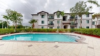 Located at 14770 SW 264th St in Homestead, Florida, this residential community offers a well-appointed outdoor amenity space designed for comfortable South Florida living. The centerpiece of the community is a generously sized resort-style swimming pool with crystal-clear blue water, complemented by a separate spa/hot tub area, all set on a spacious tiled pool deck ideal for lounging and recreation.

Adjacent to the pool is a dedicated clubhouse building, featuring a covered colonnade with striking white columns, large glass doors, and a pitched hip roof with decorative tile accents. The structure provides both functional amenities and a shaded gathering area overlooking the pool.

The community buildings are multi-story, finished in crisp white stucco with dark brown trim accents and feature private balconies on upper floors. Tall, swaying palm trees and neatly trimmed hedgerows frame the pool area, creating a sense of privacy and a distinctly tropical ambiance. Gated access points with black iron fencing help define and secure the amenity space.

The overall design reflects a Mediterranean-inspired architectural style, blending clean lines with lush greenery to create an inviting outdoor environment well-suited to the warm Homestead climate.