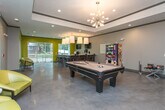 Located at 1345 Hardeman Ave in Macon, Georgia, this single-floor community amenity space offers a lively and well-appointed environment designed for socializing and recreation. The interior is defined by polished concrete floors throughout, which lend an industrial-modern character to the space.

A central gathering area serves as the heart of the room, highlighted by a full-size pool table beneath a stylish sputnik chandelier and recessed lighting with a tray ceiling detail. Adjacent to this is a substantial bar-height counter with granite countertops and metal stools, illuminated by large pendant lights — ideal for casual dining or collaborative use. Wall-mounted televisions are positioned for easy viewing from multiple areas.

Vibrant chartreuse accent walls inject energy into the space, contrasting with the neutral gray tones of the surrounding walls. Comfortable lounge seating, including distinctive yellow accent chairs, is arranged near large windows that flood the room with natural light and offer views of the lush, landscaped courtyard and surrounding greenery.

A vending machine and whiteboard add functional convenience, while direct glass-door access to an outdoor patio — equipped with a grill and shaded seating — seamlessly extends the living space outdoors. The overall ambiance is modern, social, and welcoming.