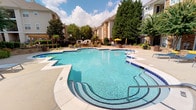 This residential community amenity space offers a thoughtfully designed environment that balances outdoor recreation with practical indoor facilities. The centerpiece of the outdoor area is a generously sized, freeform swimming pool with striking blue tile accents and a gradual entry design, making it both visually appealing and accessible. The pool deck is surrounded by mature trees and lush greenery, providing a shaded and comfortable setting for lounging. Seating areas with yellow umbrellas and patio furniture are arranged nearby, offering residents a welcoming outdoor retreat. The surrounding multi-story brick buildings frame the courtyard setting, adding a sense of community and enclosure.

Indoors, the business center is designed for productivity and comfort. The main workspace features a large white conference-style table with ergonomic seating, ideal for collaborative work or study. A built-in workstation along one wall is equipped with desktop computers and a printer, providing essential office tools. Crown molding and neutral warm-toned walls give the interior a polished, professional appearance. Light wood-style flooring runs throughout, adding warmth to the space. Large windows fitted with horizontal blinds allow natural light to filter in while maintaining privacy, with views of the surrounding greenery visible from individual workstations.