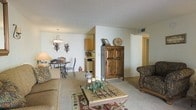 This single-floor space showcases an open-concept layout that seamlessly connects the living room, dining area, and kitchen. The living room features plush, neutral-toned carpeting and is furnished with comfortable upholstered seating arranged around a rustic wooden coffee table. A Southwestern-style area rug anchors the seating area, complementing the warm, earthy tones throughout. A prominent wooden armoire serves as a striking focal point, adding both character and storage to the space. The dining area is defined by an elegant wrought-iron chandelier and a matching bistro-style table and chairs, with decorative wall sconces adding ambient lighting. Adjacent to the dining space, the kitchen is partially visible and features wooden cabinetry with ample storage. A textured popcorn ceiling is consistent throughout, and a hallway visible from the main living area suggests access to additional rooms beyond. The overall ambiance is cozy and inviting, with a cohesive Southwestern aesthetic carried through the decor, furnishings, and accent pieces.