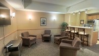 This single-floor space presents a comfortable and functional communal lounge designed for relaxed social interaction. The seating area is arranged with several plush, upholstered armchairs in a warm taupe tone, positioned to encourage conversation and ease of movement throughout the room. The walls are finished in a soft, neutral beige palette and accented with a decorative chair rail that adds a subtle architectural detail. Warm wall sconces provide a gentle, ambient glow, complementing the overall inviting atmosphere of the space. A framed black-and-white photograph serves as a tasteful focal point on the main wall, adding a classic touch to the décor. Towards one side of the room, a raised bar-height table flanked by wooden chairs offers an alternative seating option, suitable for casual dining or informal meetings. A decorative indoor tree adds a natural, refreshing element to the corner of the space. The patterned carpeted flooring ties the room together, contributing to its warm and welcoming character. Overall, the space is thoughtfully arranged to balance comfort, functionality, and a pleasant communal ambiance suitable for a variety of social activities.