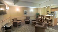 This single-floor space presents a comfortable and functional communal lounge designed for relaxed social interaction. The seating area is arranged with several plush, upholstered armchairs in a warm taupe tone, positioned to encourage conversation and ease of movement throughout the room. The walls are finished in a soft, neutral beige palette and accented with a decorative chair rail that adds a subtle architectural detail. Warm wall sconces provide a gentle, ambient glow, complementing the overall inviting atmosphere of the space. A framed black-and-white photograph serves as a tasteful focal point on the main wall, adding a classic touch to the décor. Towards one side of the room, a raised bar-height table flanked by wooden chairs offers an alternative seating option, suitable for casual dining or informal meetings. A decorative indoor tree adds a natural, refreshing element to the corner of the space. The patterned carpeted flooring ties the room together, contributing to its warm and welcoming character. Overall, the space is thoughtfully arranged to balance comfort, functionality, and a pleasant communal ambiance suitable for a variety of social activities.