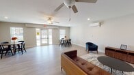 Located in Spotsylvania, VA, this well-appointed single-floor space offers a clean, modern environment designed for comfort and versatility. The open layout is anchored by light-toned hardwood-style flooring that runs throughout, complementing the neutral, warm-white walls and crisp white baseboards that give the interior a fresh, polished feel.

The main gathering area is generously proportioned, providing ample room for both lounging and dining. A seating arrangement featuring a tufted leather sofa and a pair of navy blue accent chairs surrounds a round coffee table atop a decorative area rug, creating an inviting conversation nook. Nearby, a round dining table with black chairs offers a dedicated space for meetings or meals. Recessed lighting and ceiling fans with integrated lighting ensure the space remains bright and well-ventilated throughout.

A wall-mounted ductless mini-split HVAC unit provides efficient climate control, while multiple windows dressed with horizontal blinds allow natural light to fill the room. A set of glass-paned French doors opens directly to the outdoors, further enhancing the sense of openness and providing easy exterior access with views of the surrounding greenery. The space also includes a kitchenette or utility area visible toward the rear. Overall, this space presents a clean, functional, and welcoming atmosphere suitable for a variety of uses.