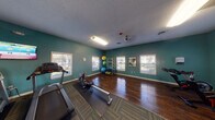 This fitness center is thoughtfully designed to accommodate a wide range of workout routines within a single-story layout. The space is divided into distinct functional zones, each serving a specific training purpose. Teal-painted walls provide a consistent, energizing aesthetic throughout, complemented by textured ceilings and a combination of dark hardwood flooring and patterned carpet tiles that delineate different activity areas.

The cardio zone features multiple treadmills, an elliptical, and a rowing machine arranged along one wall, with wall-mounted televisions positioned for convenient viewing during workouts. Full-length mirrors line an adjacent wall, visually expanding the space and supporting proper form during exercise. A cable machine and adjustable weight bench serve the strength training area, while a dedicated rack holds a comprehensive set of dumbbells.

A separate studio area with polished hardwood floors provides open space for group exercise or individual training. This zone includes stationary spin bikes, a rack stocked with resistance bands, stability balls in various sizes, and exercise mats. Multiple windows throughout the facility allow natural light to enter from several directions, creating a bright and motivating environment. An additional room accessible via interior doorway appears to provide supplementary utility space.